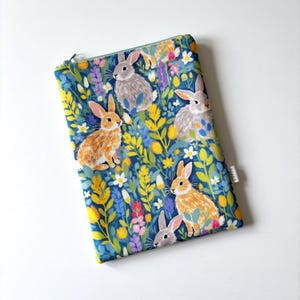 May include: A rectangular zipped pouch featuring a colourful pattern of rabbits and wildflowers. The rabbits are in shades of brown and grey, set against a dark blue background with yellow, white, and purple floral accents. The pouch has a teal zip.