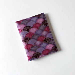 May include: A rectangular, fabric item with a repeating, fan-shaped pattern in shades of purple, magenta, and gold. The item has a dark purple border and appears to be a case or cover.