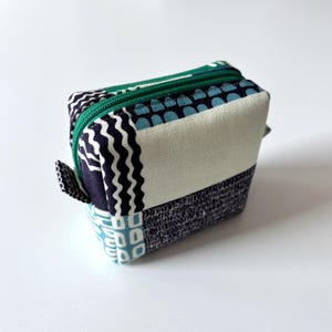 May include: A small, square fabric pouch with a green zip and pull tab. The pouch is made of various patterned fabrics in shades of blue, white, and navy. The patterns include wavy lines, geometric shapes, and a textured bottom.