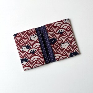 May include: A red and white fabric wallet with a blue and white polka dot interior. The exterior features a repeating pattern of stylized waves and black and white cats.