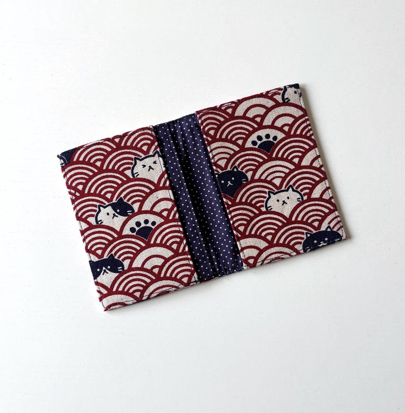 May include: A red and white fabric wallet with a blue and white polka dot interior. The exterior features a repeating pattern of stylized waves and black and white cats.