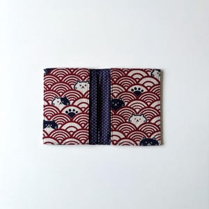 May include: A red and white fabric wallet with a repeating pattern of black cats and blue waves. The wallet has a blue and white polka dot fabric lining.