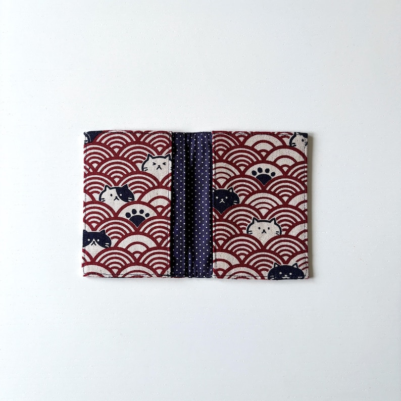May include: A red and white fabric wallet with a repeating pattern of black cats and blue waves. The wallet has a blue and white polka dot fabric lining.
