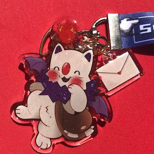 Puede incluir: Un llavero acrílico transparente con un personaje blanco con forma de gato con alas de murciélago moradas, sosteniendo una bolsa marrón. El llavero incluye un pequeño dije de sobre con un corazón y un cordón azul con texto blanco. El fondo es rojo.