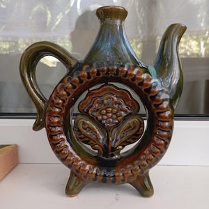 以下が含まれることがあります： ユニークなデザインの茶色と緑色のセラミックピッチャー。中央に花の模様がある円形の開口部、ハンドル、注ぎ口があります。ピッチャーは3つの小さな足の上にあり、光沢のある釉薬仕上げです。装飾的なアイテムです。
