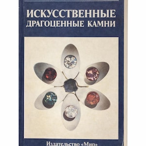 May include: A vintage book titled "Artificial Gems" in Russian. The cover features a flower-like design with various gemstones in different colors. The book has a dark blue spine and cover edges, with text in white.
