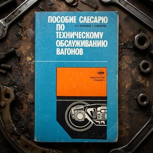 Puede incluir: Un manual técnico azul titulado POSOBIE SLESARYU PO TEKHNICHESKOMU OBSLUZHIVANIYU VAGONOV con un gráfico naranja y negro de un conjunto de ruedas de tren en la portada, con texto cirílico blanco y el nombre de la editorial TRANSPORT.