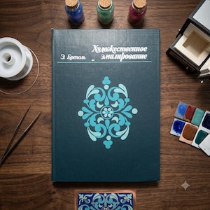 May include: A dark teal hardcover book titled "Художественное эмалирование" by Э. Бреполь, featuring a light blue symmetrical floral pattern on the cover. The book is surrounded by three small glass jars, a spool of wire, tweezers, and a paint palette.