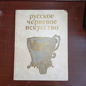 May include: A vintage book with the title "русское черневое искусство" in gold lettering. The cover features a gold and gray ornate cup design. The book is on a dark wood surface.