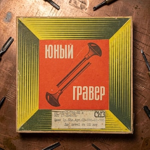 May include: A square cardboard box with a red center panel featuring a black graphic of a tool and the Cyrillic text "ЮНЫЙ ГРАВЕР". The border is yellow with green diagonal lines. A white label at the bottom contains technical text and pricing.