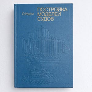 Op de afbeelding: Een blauw boek met gouden letters en reliëf illustraties van zeilschepen. De titel, in goud, luidt "Constructie van scheepsmodellen" in het Russisch. Het boek staat tegen een witte achtergrond.