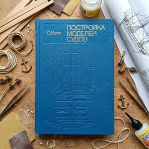 May include: A blue hardback book titled ПОСТРОЙКА МОДЕЛЕЙ СУДОВ by O. Kurti, featuring an embossed line drawing of a sailing ship on the front cover. The book is surrounded by wooden strips, rope coils, small metal anchors, tweezers, and a glue bottle.