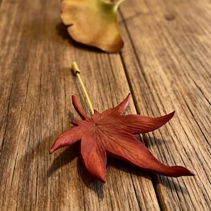 May include: A red maple leaf lapel pin featuring a detailed, textured surface and a thin gold-toned metal needle. The leaf has five distinct, pointed lobes with visible veins, and the pin is positioned upright with the needle extending vertically.