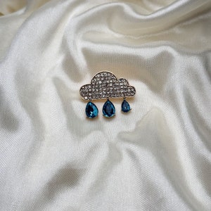 Puede incluir: Un broche de tono dorado con forma de nube con gotas de lluvia de piedras preciosas azules.