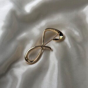 May include: A gold infinity symbol brooch. The brooch is made of a smooth, shiny metal and has a simple, elegant design.
