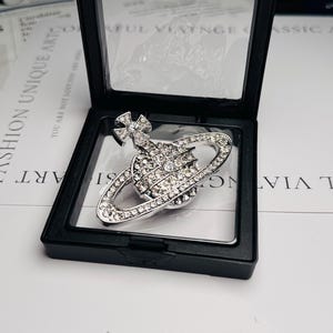 Puede incluir: Broche plateado con forma de planeta, adornado con cristales transparentes y una cruz. Se muestra en una caja negra con tapa transparente, sobre un fondo blanco.