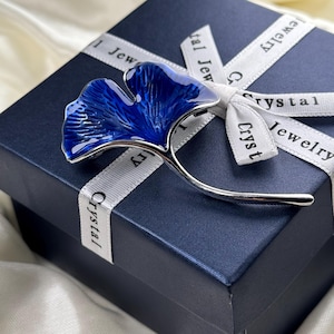 Peut inclure: Une broche en forme de feuille de ginkgo bleue avec des accents argentés, présentée sur un coffret à bijoux bleu avec un ruban blanc qui porte l'inscription "Crystal Jewelry".