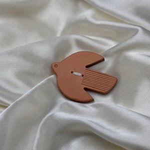 May include: A terracotta-colored ceramic dove ornament with a white string attached to it. The dove is sitting on a white satin fabric.