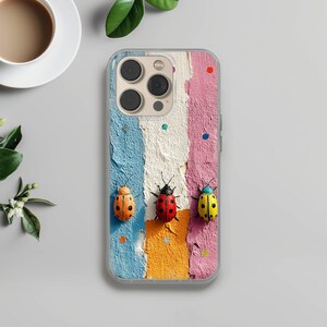 Könnte beinhalten: Transparente Handyhülle mit einem strukturierten Design aus vertikalen Streifen in Blau, Weiß, Rosa und Orange. Die Hülle zeigt drei Marienkäferfiguren in Orange, Rot und Gelb. Die Hülle liegt auf einer grauen Oberfläche mit einer Tasse Kaffee.
