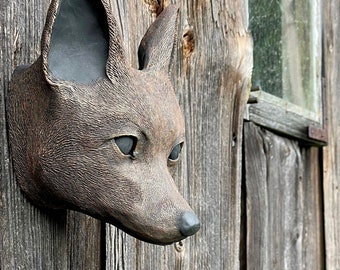 May include: A wall-mounted sculpture of a fox head with a textured bronze-toned finish. The fox features pointed ears with dark inner detailing, almond-shaped eyes, and a smooth, dark-colored nose, projecting outward from the mounting surface.
