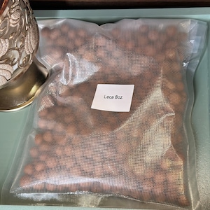 May include: A clear plastic bag filled with brown, spherical leca clay pebbles. A white label on the bag reads "Leca 8oz.". The bag is sealed and sits on a light green tray.