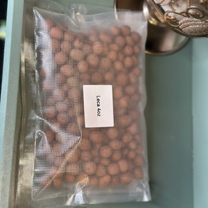 May include: A clear plastic bag filled with small, brown, round clay pellets. The bag is labeled "Leca 4oz".