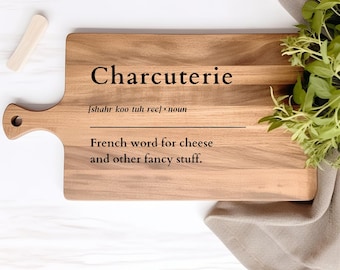 Può includere: Un tagliere di legno con manico, che presenta la parola "Charcuterie" e la sua definizione: "[shahr koo tuh reef] sostantivo Parola francese per formaggio e altre cose raffinate."
