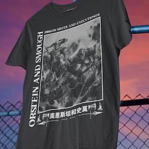 May include: Black t-shirt featuring a grayscale graphic of a battle scene with a dragon and knight. The shirt has the text "ORSTEIN AND SMOUGH" vertically on the left and "DRAGON SLAYER AND EXECUTIONER" at the top.