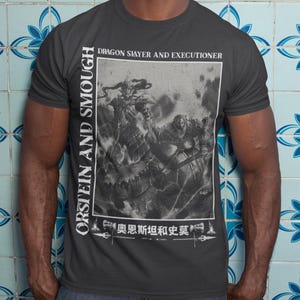 May include: Dark grey t-shirt with a black and white graphic depicting a battle scene, with the text "DRAGON SLAYER AND EXECUTIONER." The shirt also features the text "ORSTEIN AND SMOUGH" vertically and Japanese text at the bottom.
