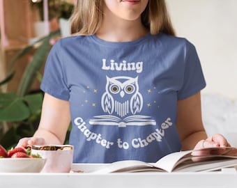 May include: A blue t-shirt with the text "Living Chapter to Chapter" and a white owl design. The owl is perched on an open book, with stars around it. The shirt is being worn.