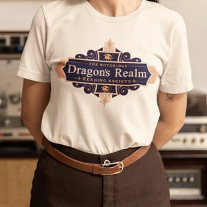 May include: A white t-shirt with a brown leather belt and brown pants. The shirt features a blue and gold design with the text "The Notorious Dragon's Realm Reading Society."