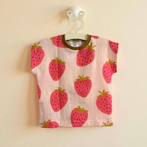 Puede incluir: Una camiseta rosa claro de manga corta con un estampado repetido de fresas grandes de color rosa brillante. La camiseta tiene un cuello marrón y está colgada de una percha blanca. Las fresas tienen tallos y semillas de color amarillo verdoso.