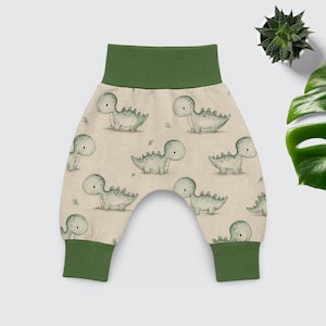 Könnte beinhalten: Babyhose mit einem sich wiederholenden Muster aus grünen Dinosauriern auf hellbraunem Hintergrund. Die Bündchen und der Bund sind einfarbig grün. Die Hose ist aus weichem Material.