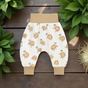 May include: Baby pants with a white background and a pattern of turtles and rainbows in shades of brown and tan. The waistband and cuffs are solid tan. The pants are made of soft fabric.