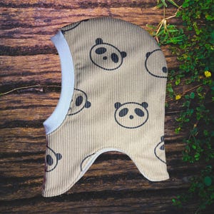 Op de afbeelding: Een bruin corduroy bivakmuts met een herhaald patroon van zwarte panda gezichten. De bivakmuts heeft een witte rand rond de gezichtsopening en is ontworpen om het hoofd en de nek te bedekken.