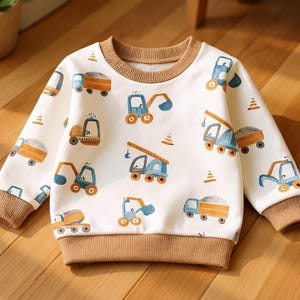 Könnte beinhalten: Weißes Sweatshirt mit Rundhalsausschnitt und einem sich wiederholenden Muster aus blauen, orangefarbenen und braunen Baufahrzeugen. Das Sweatshirt hat braune gerippte Bündchen, einen Ausschnitt und einen Saum. Baukonen sind ebenfalls auf dem Stoff verstreut.