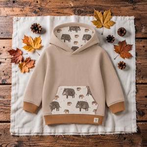 Könnte beinhalten: Ein beigefarbener Kapuzenpullover mit langen Ärmeln, gerippter Textur, Kängurutasche und einem Kapuzenfutter mit einem wiederkehrenden Muster aus braunen Wildschweinen. Die Ärmelbündchen und der untere Saum sind in einem einfarbigen Hellbraun.