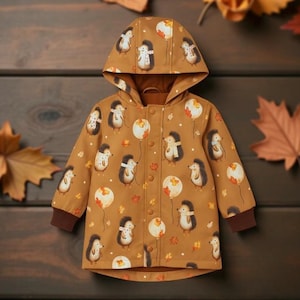 Op de afbeelding: Een bruin jasje met capuchon en een patroon van egels en ballonnen. De jas heeft een bruine voering, manchetten en knopen. Het ontwerp toont cartoon egels met ballonnen en herfstbladeren.