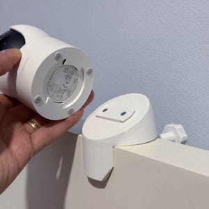 Könnte beinhalten: Eine weiße Xiaomi-Überwachungskamera mit schwarzem Objektivgehäuse und einem runden Sockel mit vier kleinen Gummifüßen. Daneben befindet sich eine weiße, maßgefertigte Halterung mit flacher Oberseite und einem Schraubmechanismus zur Befestigung.