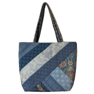 May include: A tote bag constructed from various blue fabric panels featuring a mix of quilted textures, floral damask patterns, and geometric weaves. The bag includes two long shoulder straps made from the same blue patterned fabric as the body.