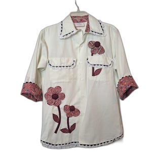 Puede incluir: Camisa crema con botones y detalles de apliques florales. Cuenta con mangas tres cuartos con puños estampados, dos bolsillos en el pecho y un ribete de encaje en el cuello, los bolsillos y el dobladillo. La camisa tiene una estética vintage y hecha a mano.