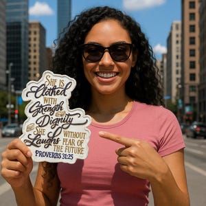May include: A decorative sign with the text "She is clothed with strength & dignity, she laughs without fear of the future, Proverbs 31:25". The sign has a cream background with navy and gold lettering.