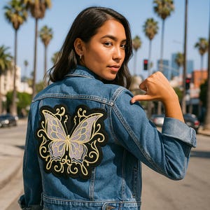 May include: A denim jacket with a large butterfly patch on the back. The patch features a butterfly with lavender and yellow details, outlined in black. The jacket is a medium wash blue and has a classic design.