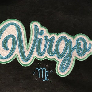 Op de afbeelding: Een zwarte stoffen patch met het woord "Virgo" in een glinsterend blauw en groen lettertype. Het astrologische symbool van de Maagd staat onder het woord in blauw.