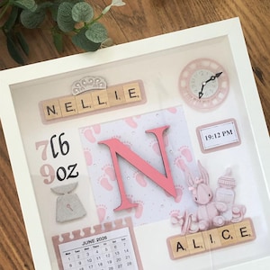 May include: A white shadow box frame containing a personalised baby keepsake with the names Nellie and Alice spelled out in wooden letter tiles, a large pink letter N, a clock showing 19:12 PM, a calendar for June 2026, and baby motifs.