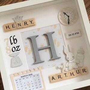 May include: A white shadow box frame containing a personalised baby keepsake display. It features the names HENRY and ARTHUR in wooden letter tiles, a large grey letter H, a clock set to 10:32 PM, a March 2026 calendar, and a baby bottle.