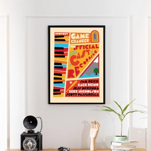 Könnte beinhalten: Ein gerahmtes Poster mit dem Text "DROPOUT GAME CHANGER THE OFFICIAL CAST RECORDING" in einem farbenfrohen Design. Das Poster enthält Namen und eine Klaviertastatur-Illustration. Das Poster wird über einer Holzkommode angezeigt.