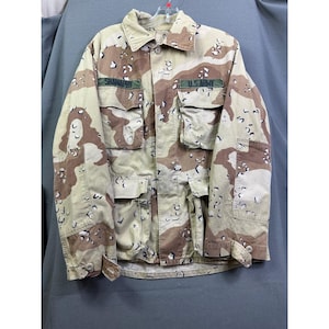 May include: A long-sleeve military-style jacket featuring a desert camouflage pattern in shades of tan, brown, and cream. The jacket includes four front pockets with button flaps, a collared neckline, and patches reading SHANAPHY and U.S. ARMY.