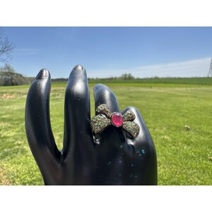 Puede incluir: Un anillo en forma de lazo con una gema rosa ovalada grande en el centro. El lazo está incrustado con pequeñas gemas verdes. El anillo se muestra en una mano negra sobre un fondo de hierba verde y cielo azul.