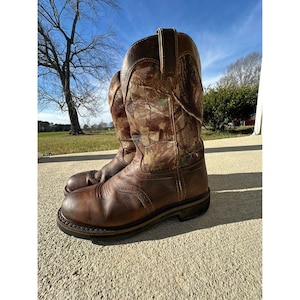 May include: A pair of brown leather cowboy boots with a camouflage pattern on the upper shaft. The boots have a rounded toe, a sturdy sole, and a pull-on design. The boots are sitting on a concrete surface.